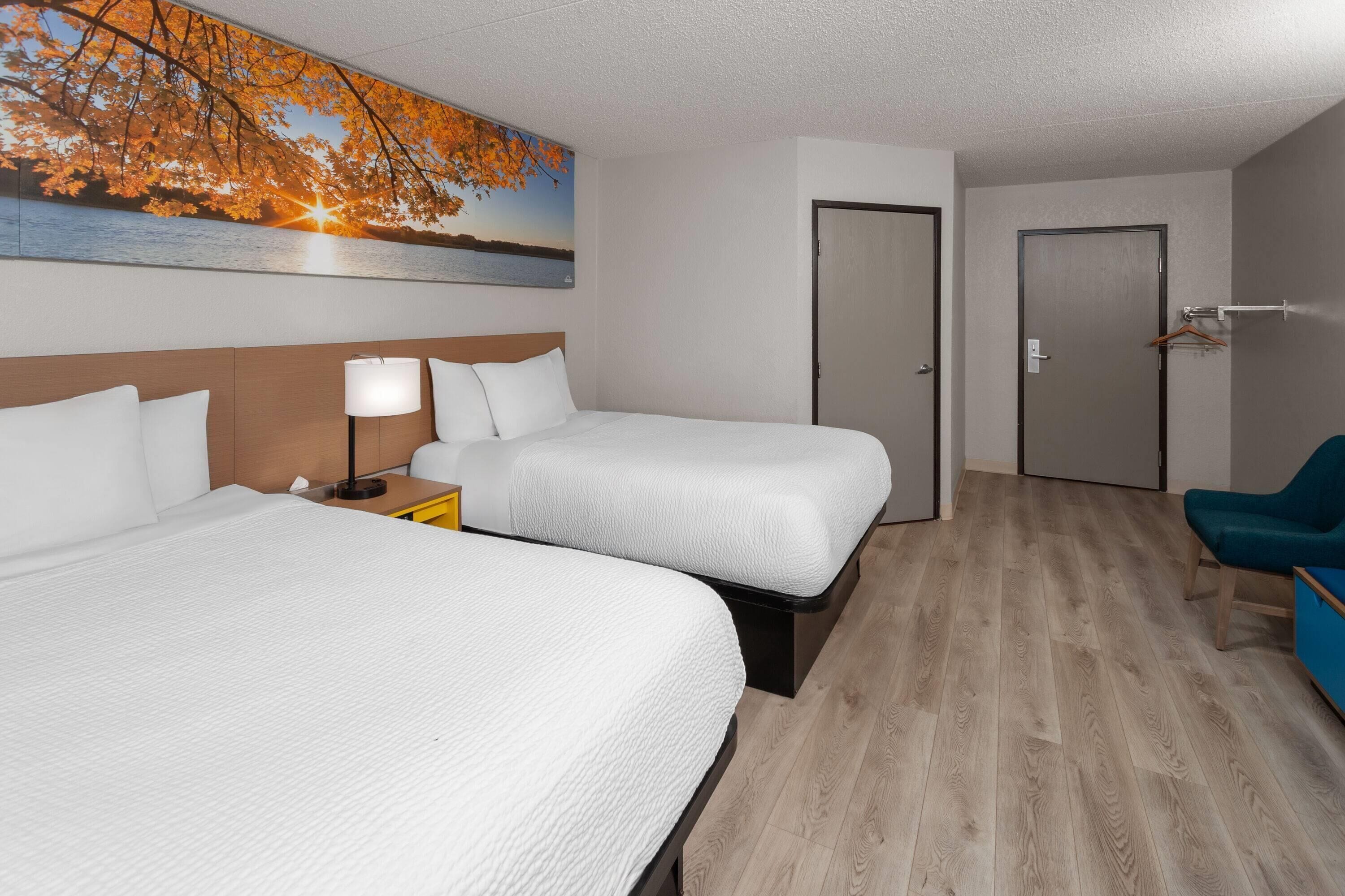 A hotel room with two beds, a painting of a sunset over water, a wooden floor, and a blue chair.