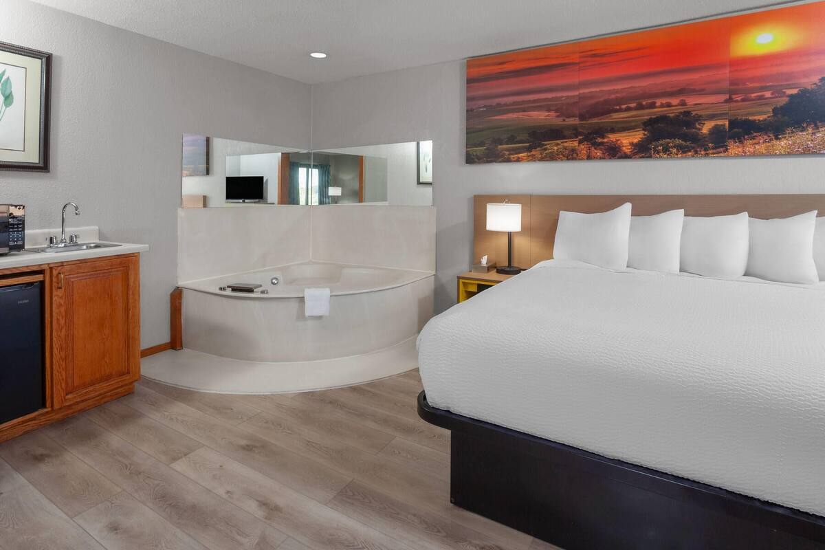 Une chambre d’hôtel avec un grand lit, une baignoire, une coiffeuse avec un lavabo et un tableau représentant un coucher de soleil accroché au mur.