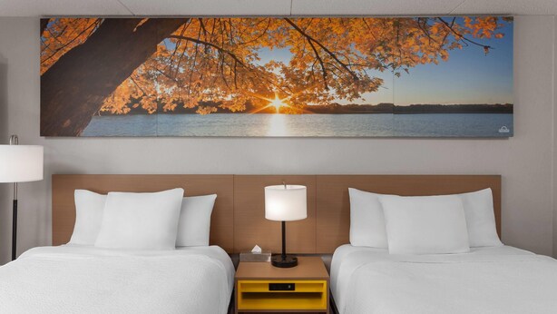 A hotel room with two beds, a nightstand with a lamp, and a large painting of a sunset over a lake. - Days Inn by Wyndham Mason City (Mason City)