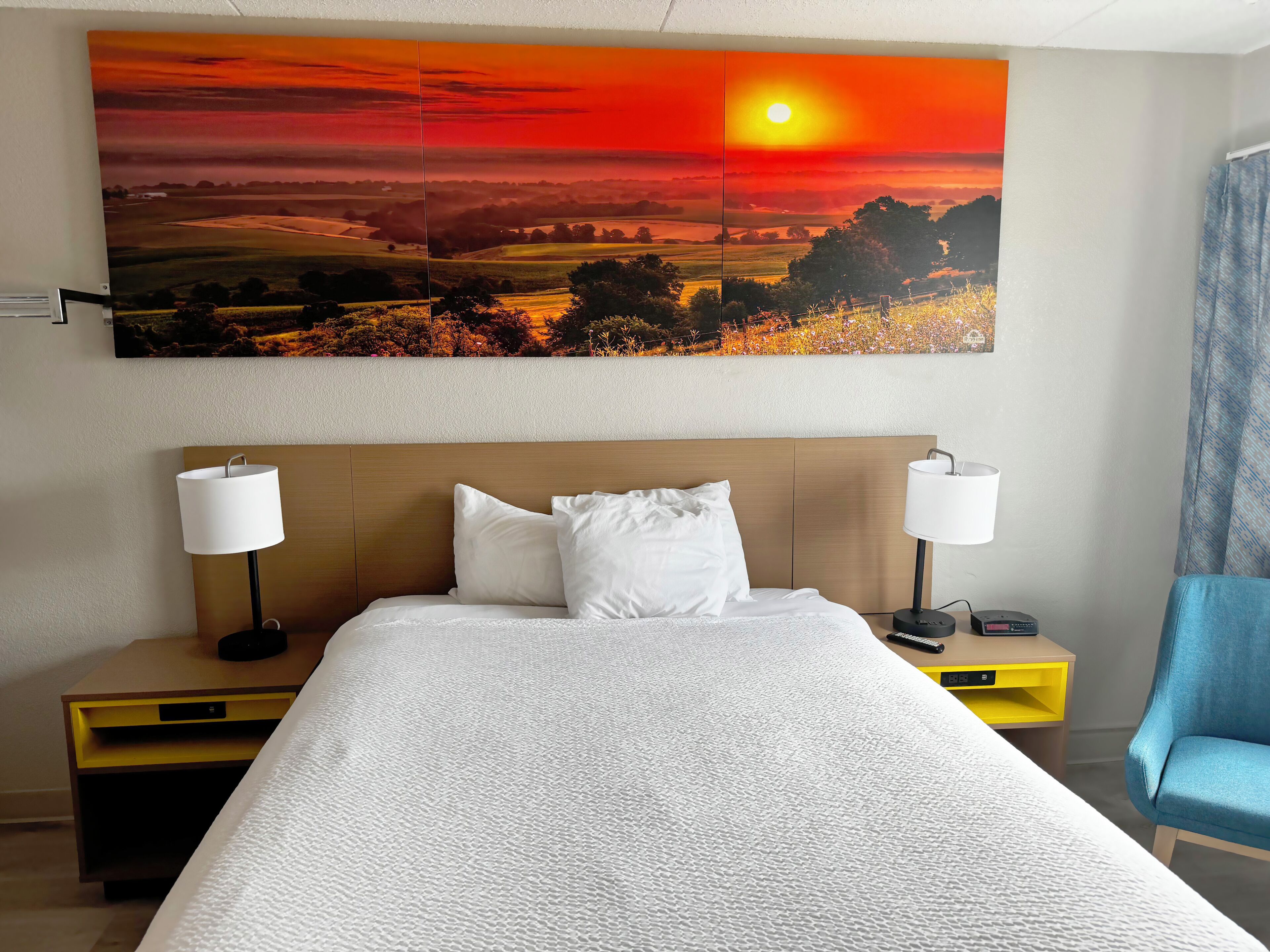 A hotel room with a large painting of a sunset, a bed with white bedding, two bedside lamps, a nightstand, and a blue armchair.