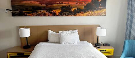 A hotel room with a large painting of a sunset, a bed with white bedding, two bedside lamps, a nightstand, and a blue armchair.