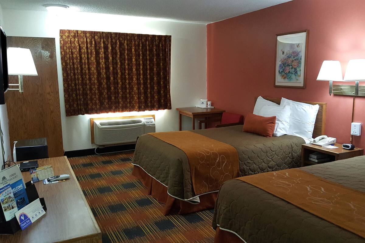 A hotel room with two beds, a desk with a lamp, a small table with a phone, and a wall-mounted air conditioner.