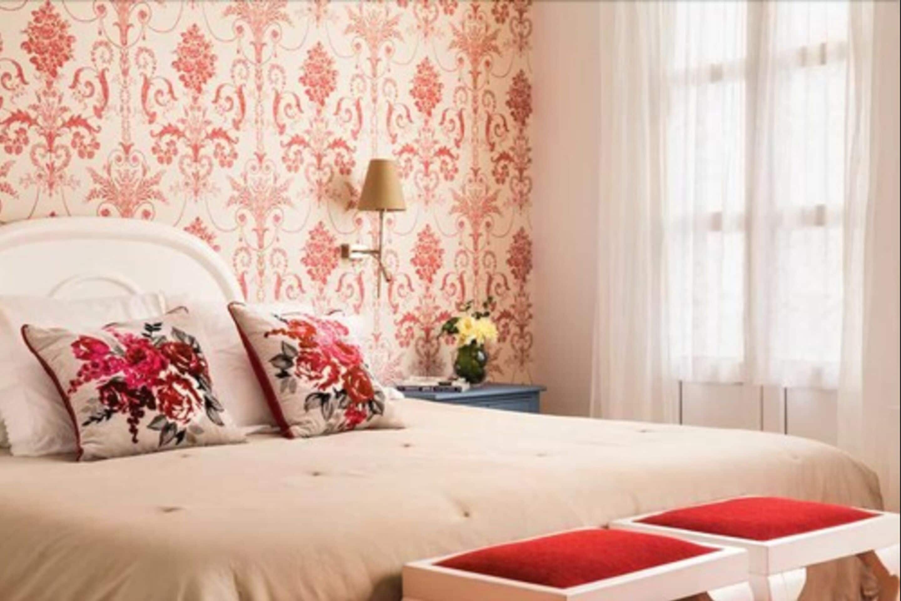 Un dormitorio con una cama, almohadillas florales, una mesita de noche con lámpara y un jarrón con flores.
