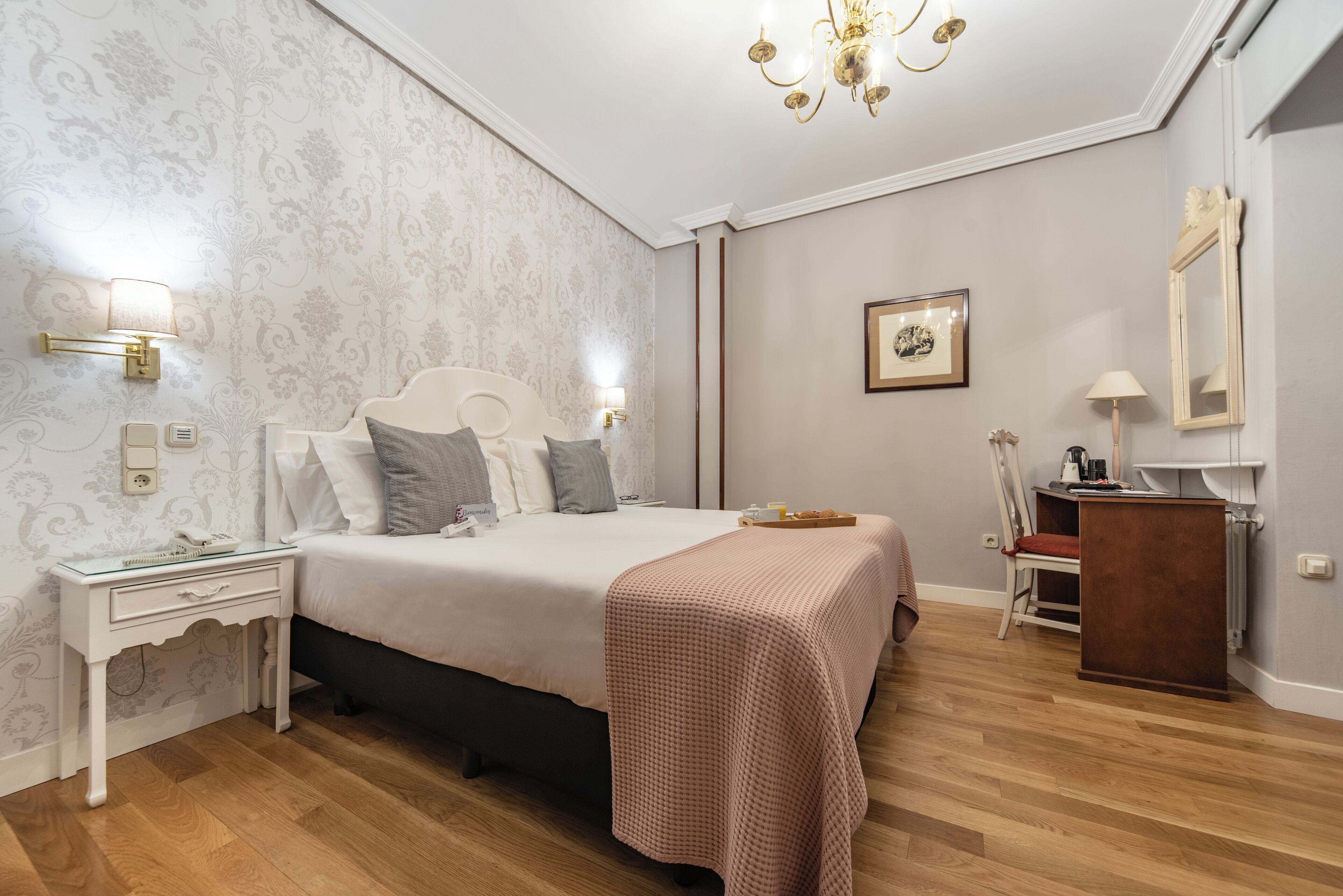 Un dormitorio con una cama grande, mesitas de noche, un tocador, una cómoda, una silla y una araña.