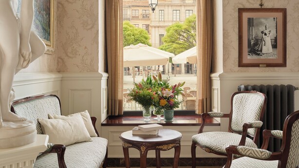 Una habitación con vistas a un patio, una estatua, una mesa con un florero y un cuadro en la pared.