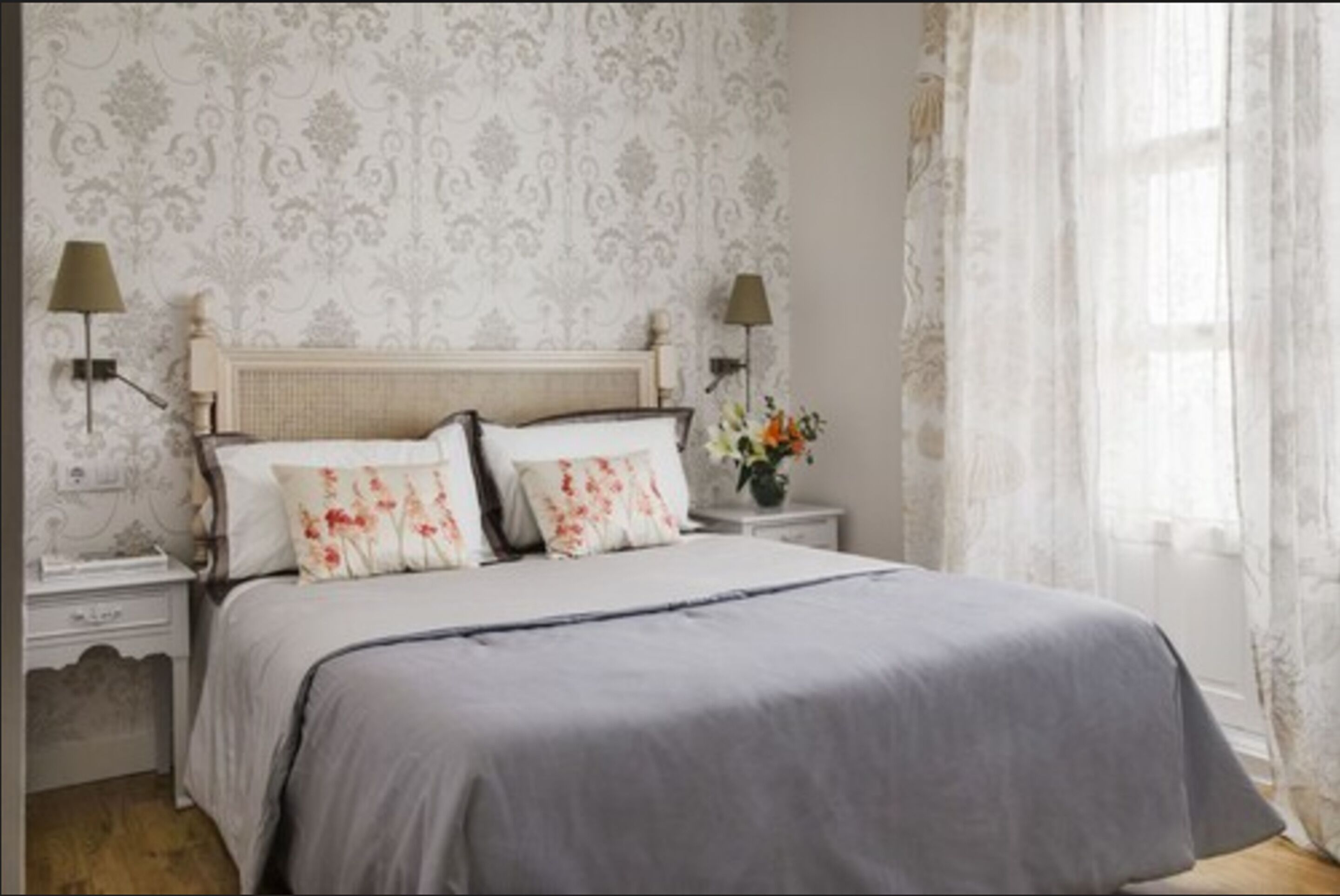 A bedroom with a large bed, two bedside tables, a nightstand, and a window with sheer curtains.