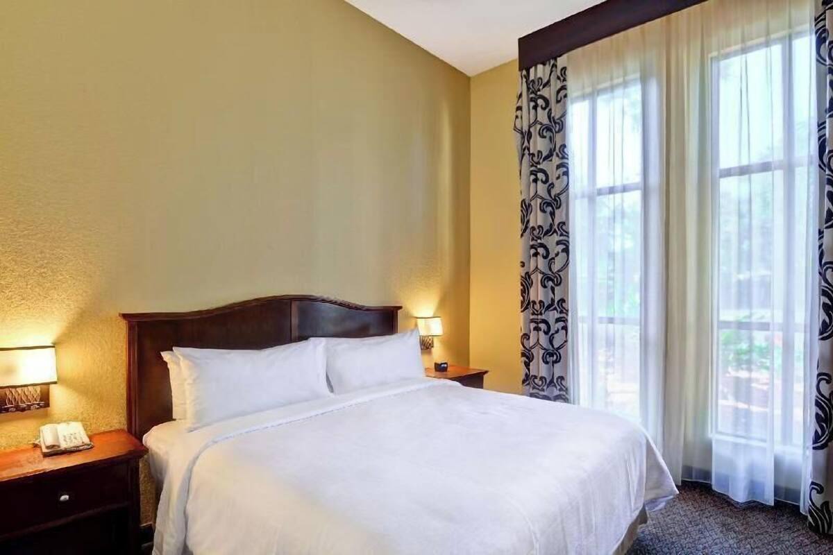 Een hotelkamer met een groot bed, een nachtkastje met een lamp en een raam met gordijnen.