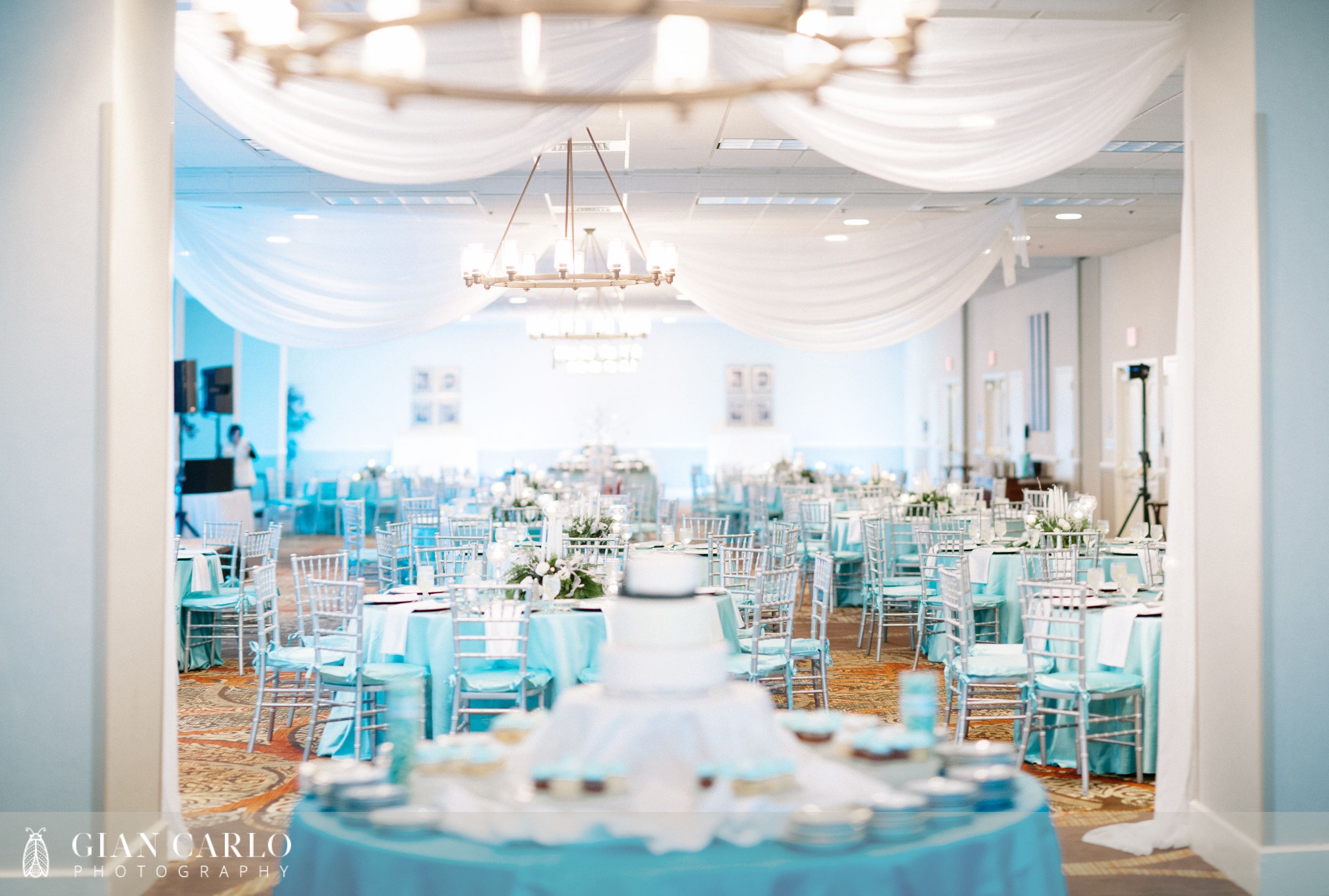 Un salón de banquetes con mesas redondas, sillas blancas y una exhibición central de pasteles.
