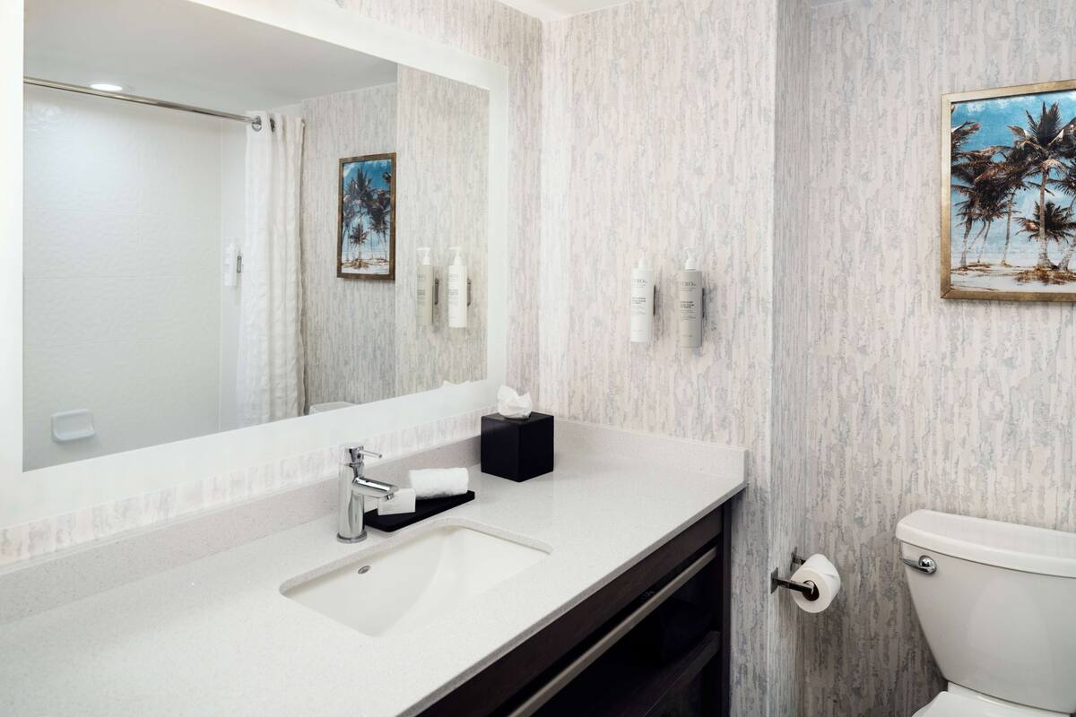 Een badkamer met een grote spiegel, een wastafel, toilet en een lijst met een afbeelding van palmbomen aan de muur.