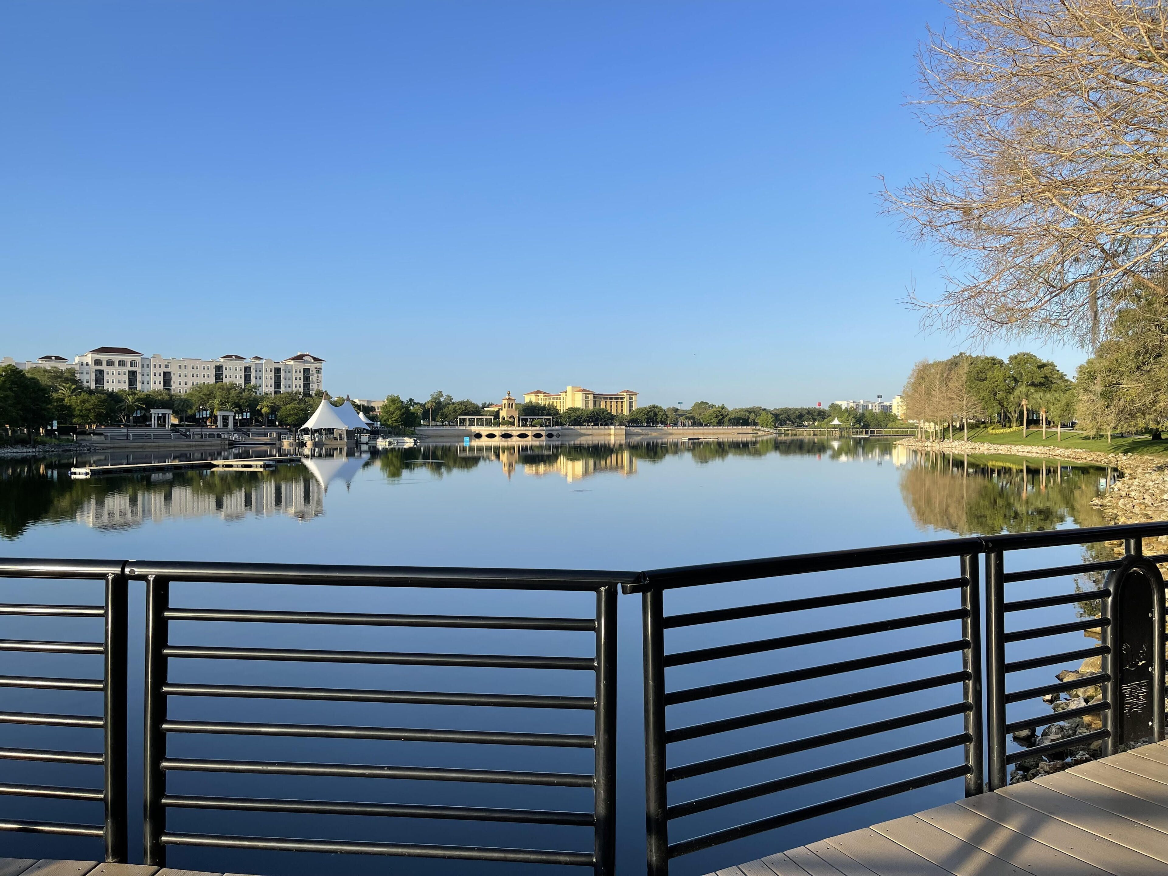 A calm lake with a metal railing in the foreground, buildings in the background, and a clear blue sky.