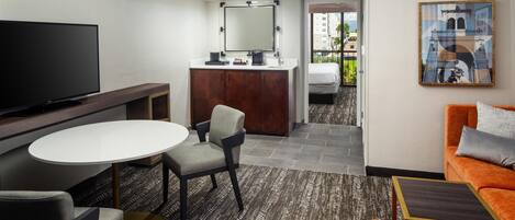 A hotel room with a flat-screen TV, a round dining table, a sofa, a chair, a small kitchenette, and a painting on the wall.