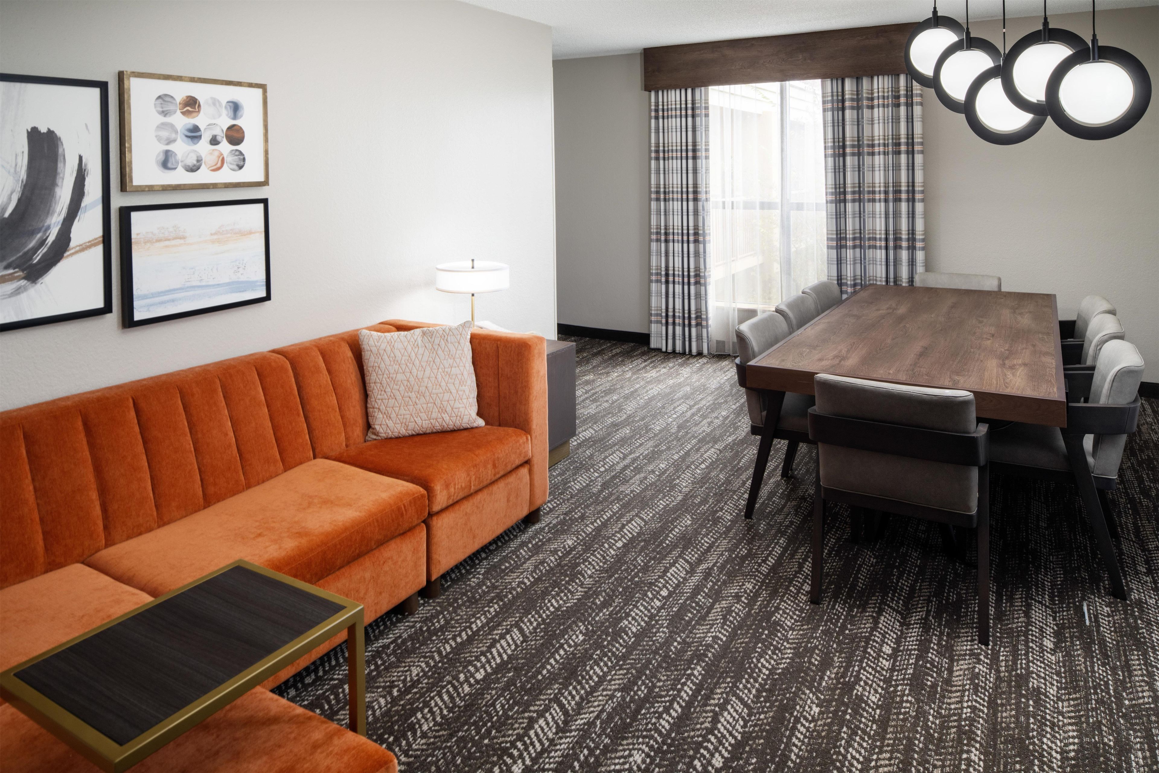 A modern hotel room with a sofa, a small table, a dining table with chairs, a lamp, and a patterned carpet.