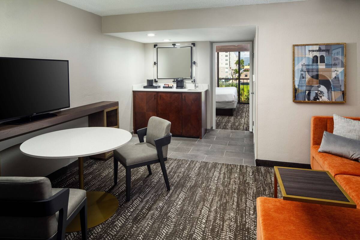 Een hotelkamer met een flatscreen televisie, een ronde eettafel, een bank, een stoel, een klein tafeltje, een kast, een spiegel en een schilderij aan de muur.