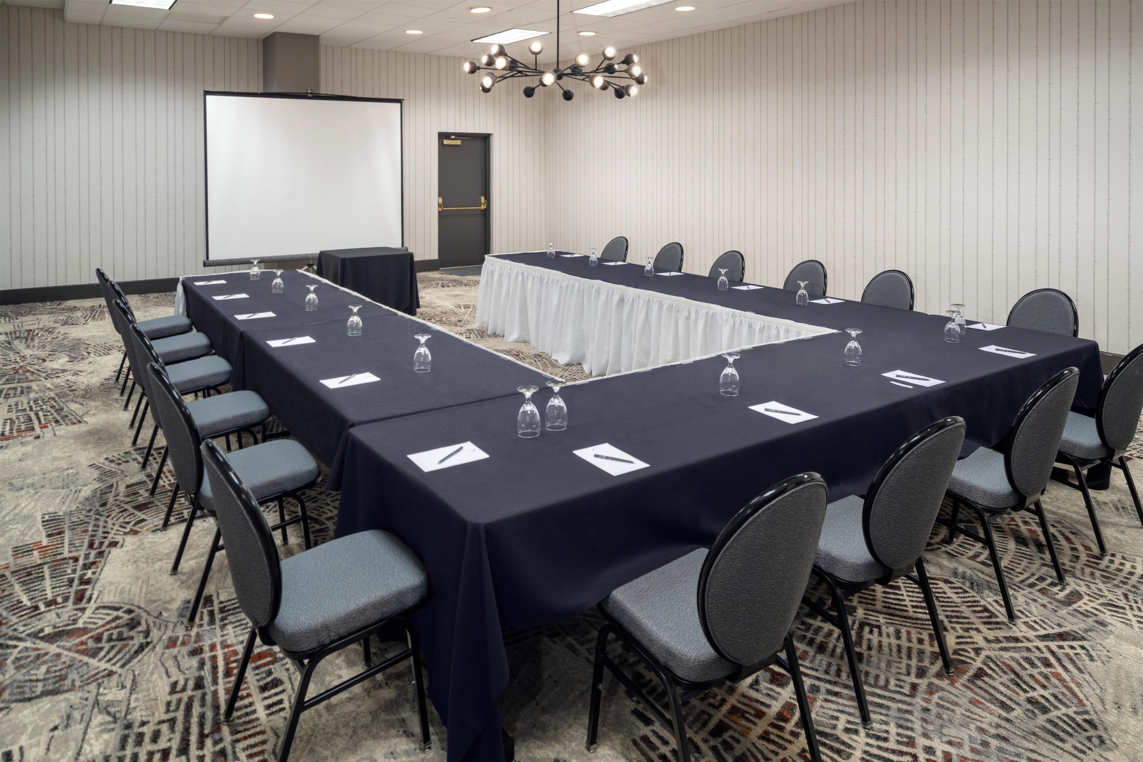 A conference room with a long table, chairs, a projector screen, and a carpeted floor.