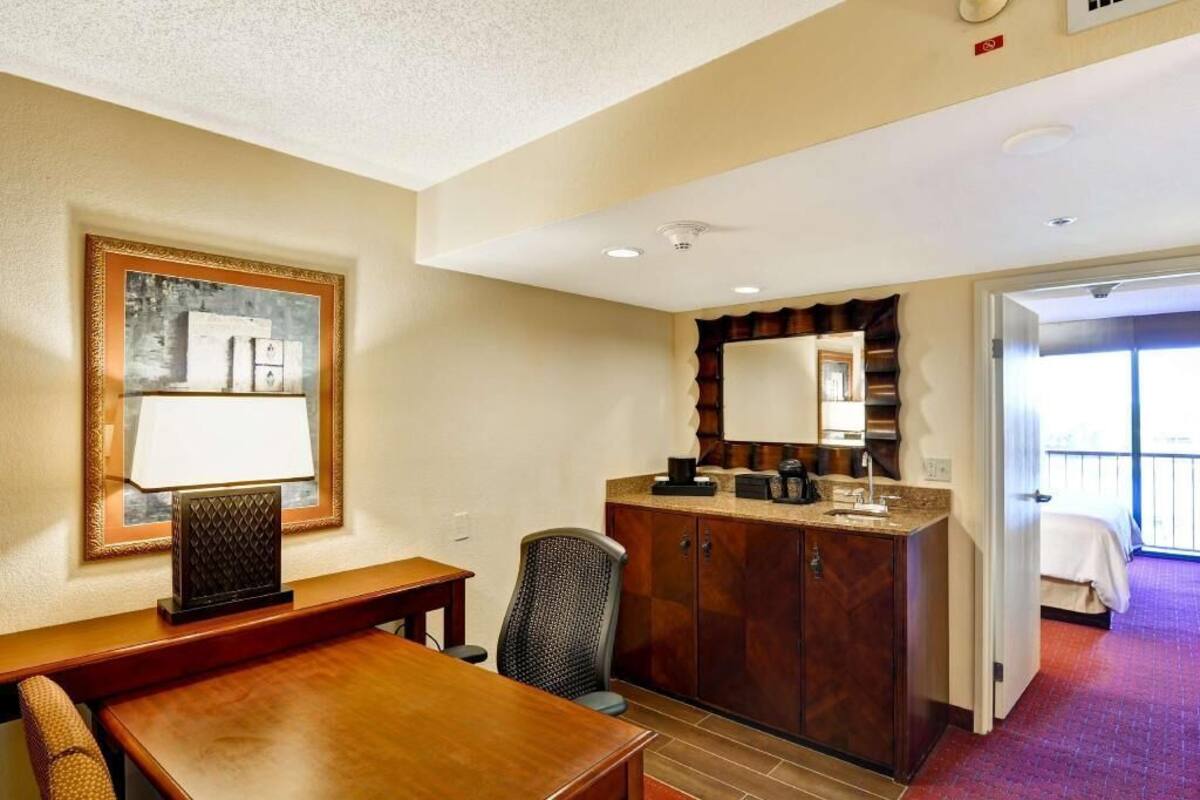Hotelkamer met een houten bureau, een stoel, een minibar, een grote spiegel en een deur naar een balkon.