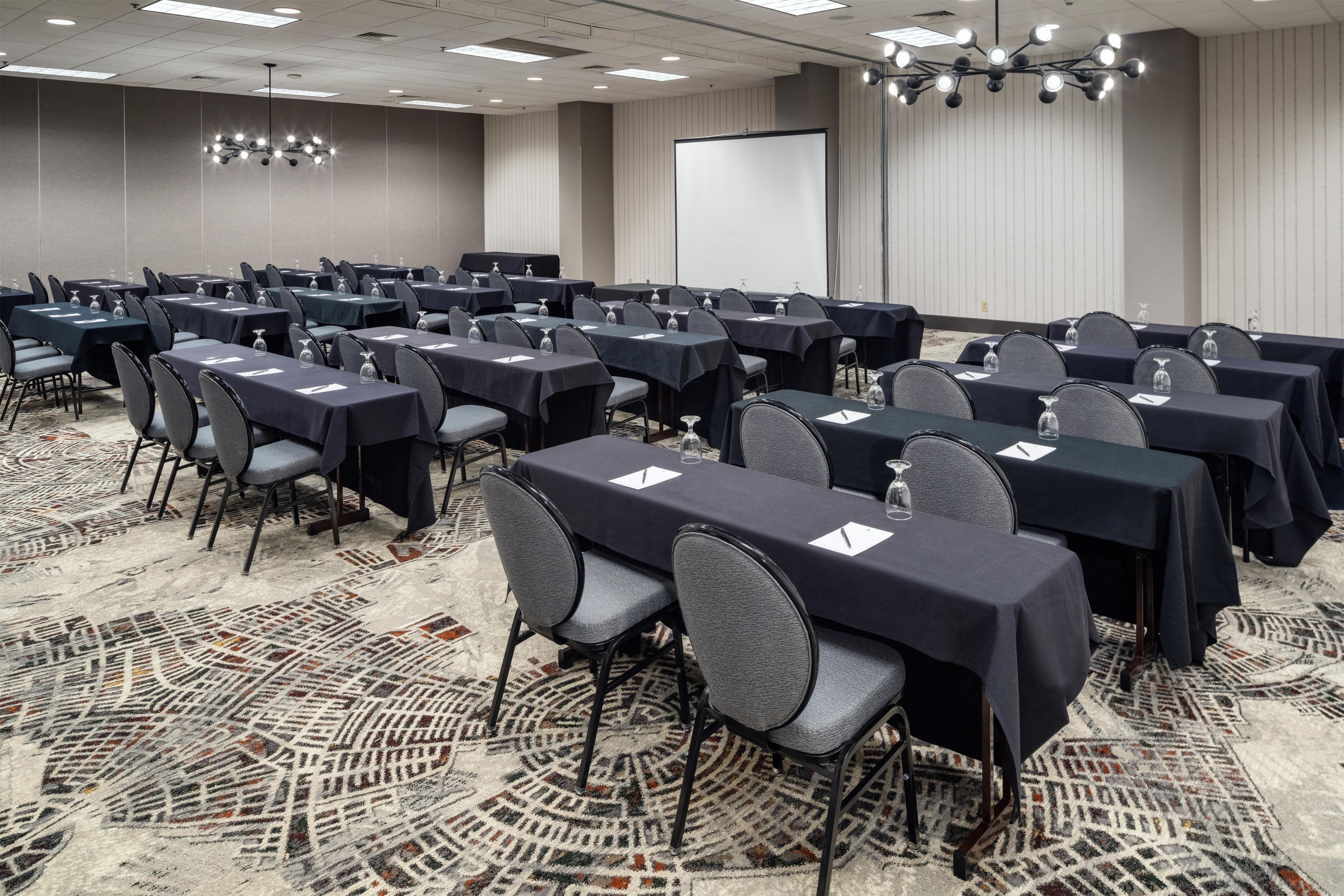 A conference room with rows of tables and chairs, a projector screen, and a patterned carpet.