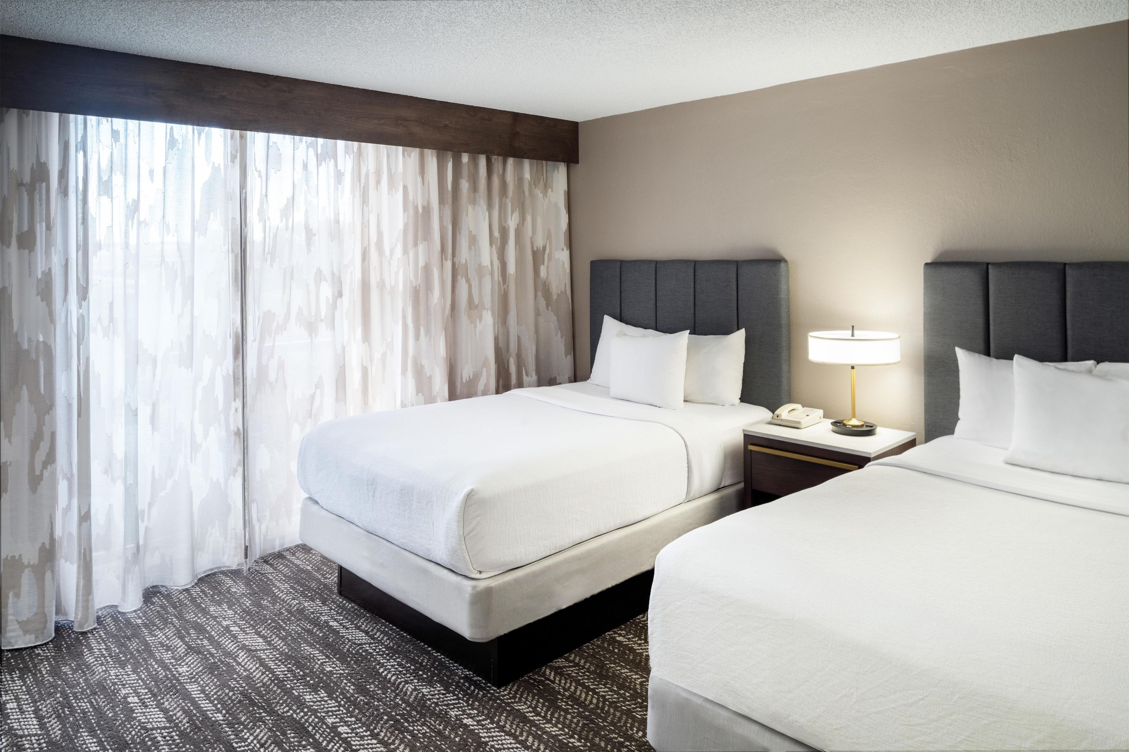 A hotel room with two beds, a nightstand with a lamp, and a large window with curtains.