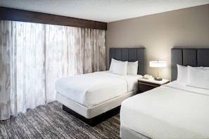 A hotel room with two beds, a nightstand with a lamp, and a large window with curtains.
