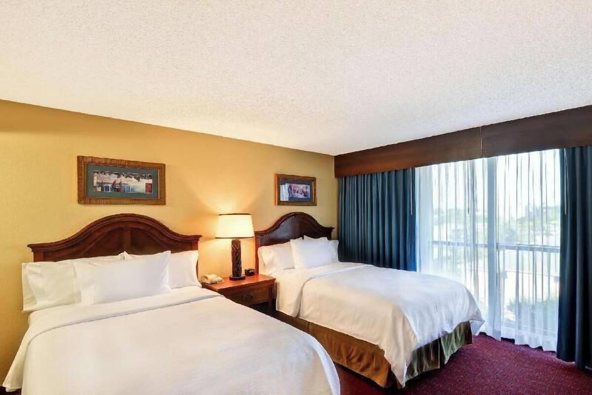 Een hotelkamer met twee bedden, een nachtkastje met een lamp en een groot raam met gordijnen.