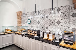 Buffet de café da manhã com diversos alimentos, placa de padaria e máquina de café.