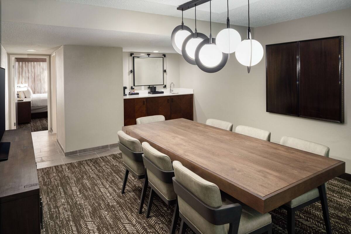 Een hotelkamer met een grote houten eettafel, vier stoelen, een kleine kitchenette, een grote spiegel en een donker houten paneel aan de muur.