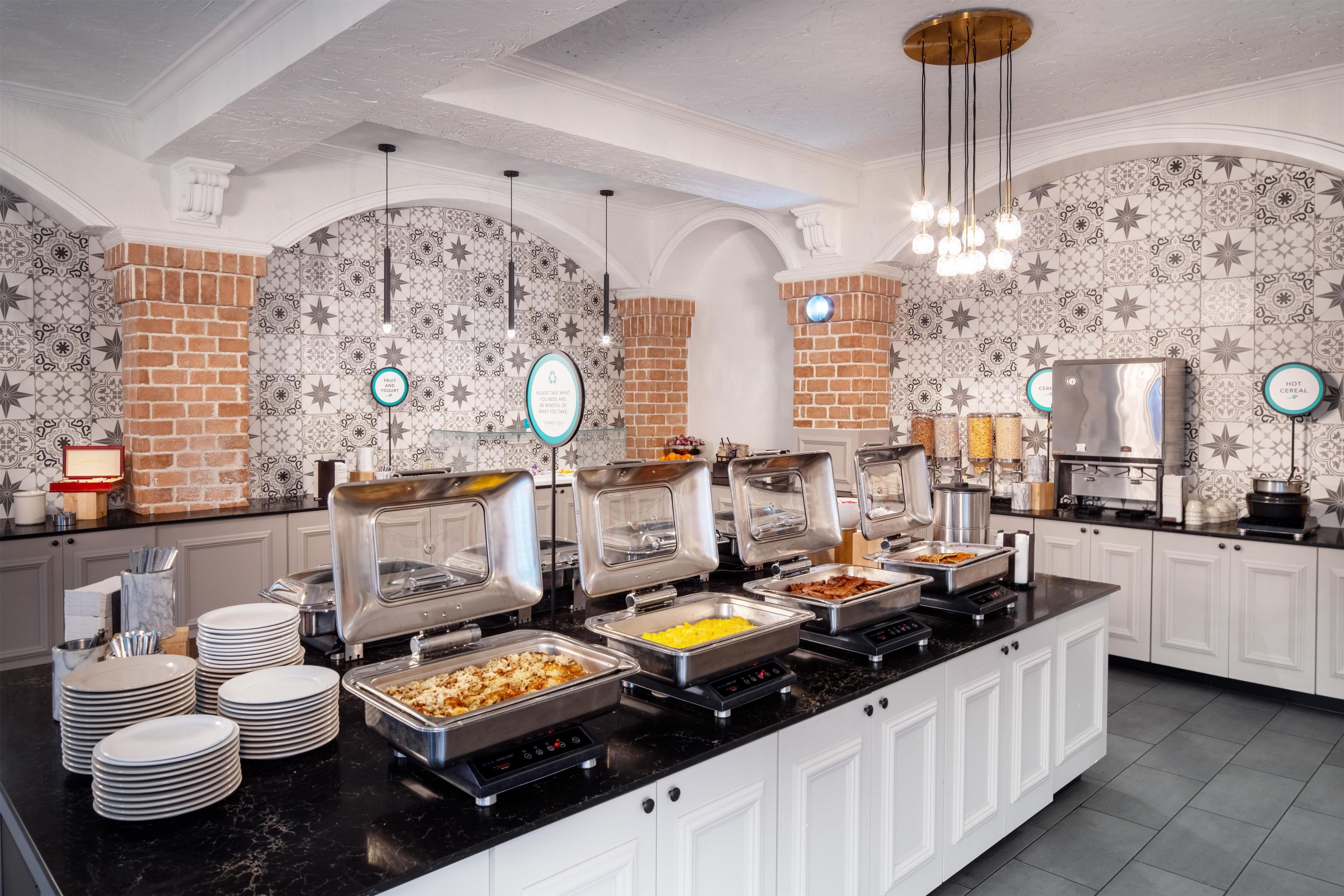A buffet setup with various food stations, including a yogurt station and a cereal station, in a room with patterned walls and a brick accent wall.
