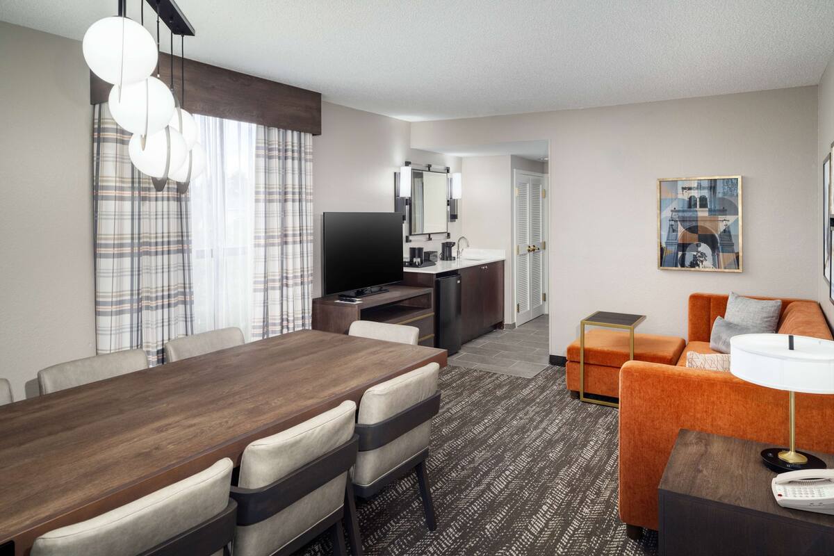 Een moderne hotelkamer met een eethoek, een bank, een televisie, een kitchenette en een schilderij aan de muur.
