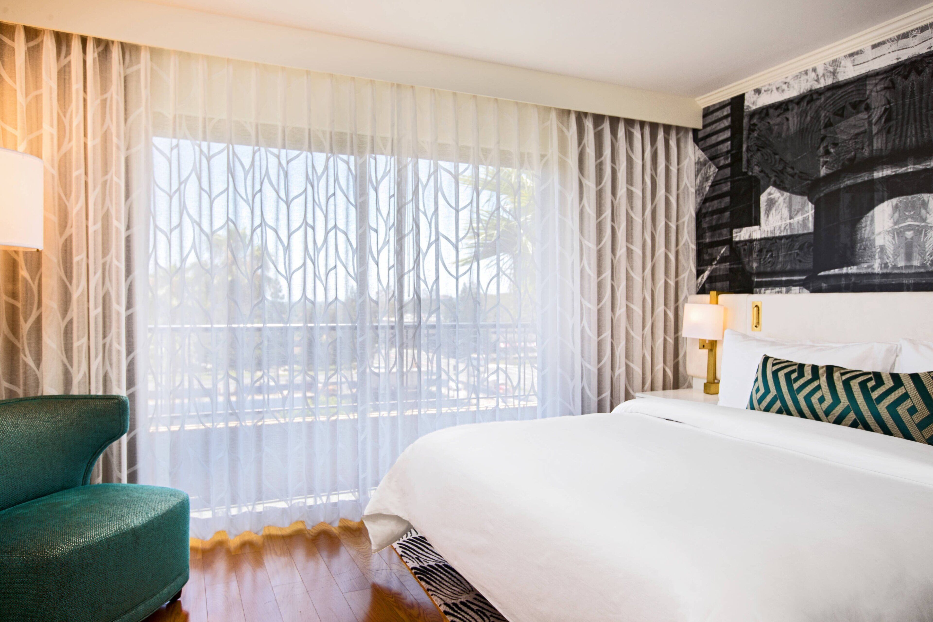 Una habitación de hotel con una cama grande, un sillón verde y un ventanal con cortinas traslúcidas.