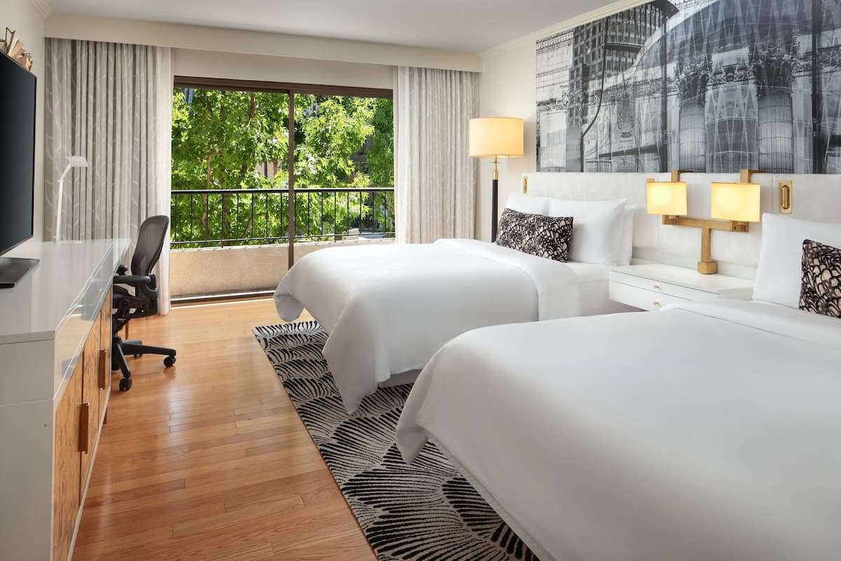 A hotel room with two beds, a desk with a chair, a TV, and a balcony with a view of trees.