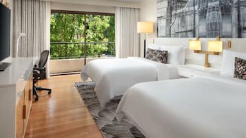 Habitación de hotel con dos camas, un escritorio con silla, televisor y balcón con vista a árboles.