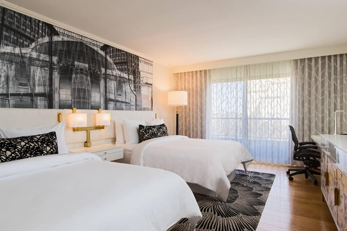 A hotel room with two beds, a desk, a chair, a lamp, and a large abstract painting on the wall.