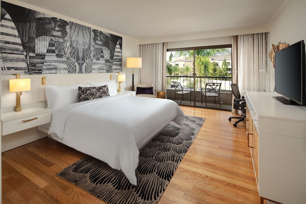 A modern hotel room with a large bed, a desk with a chair, a TV, and a balcony with a view of greenery.