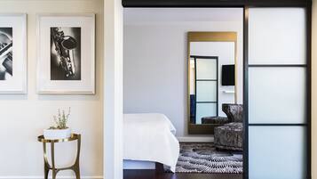 Una habitación de hotel moderna con cama, una mesita de noche con una planta, fotografías blanco y negro enmarcadas, un tapete estampado y una puerta corredera que da a otra habitación.