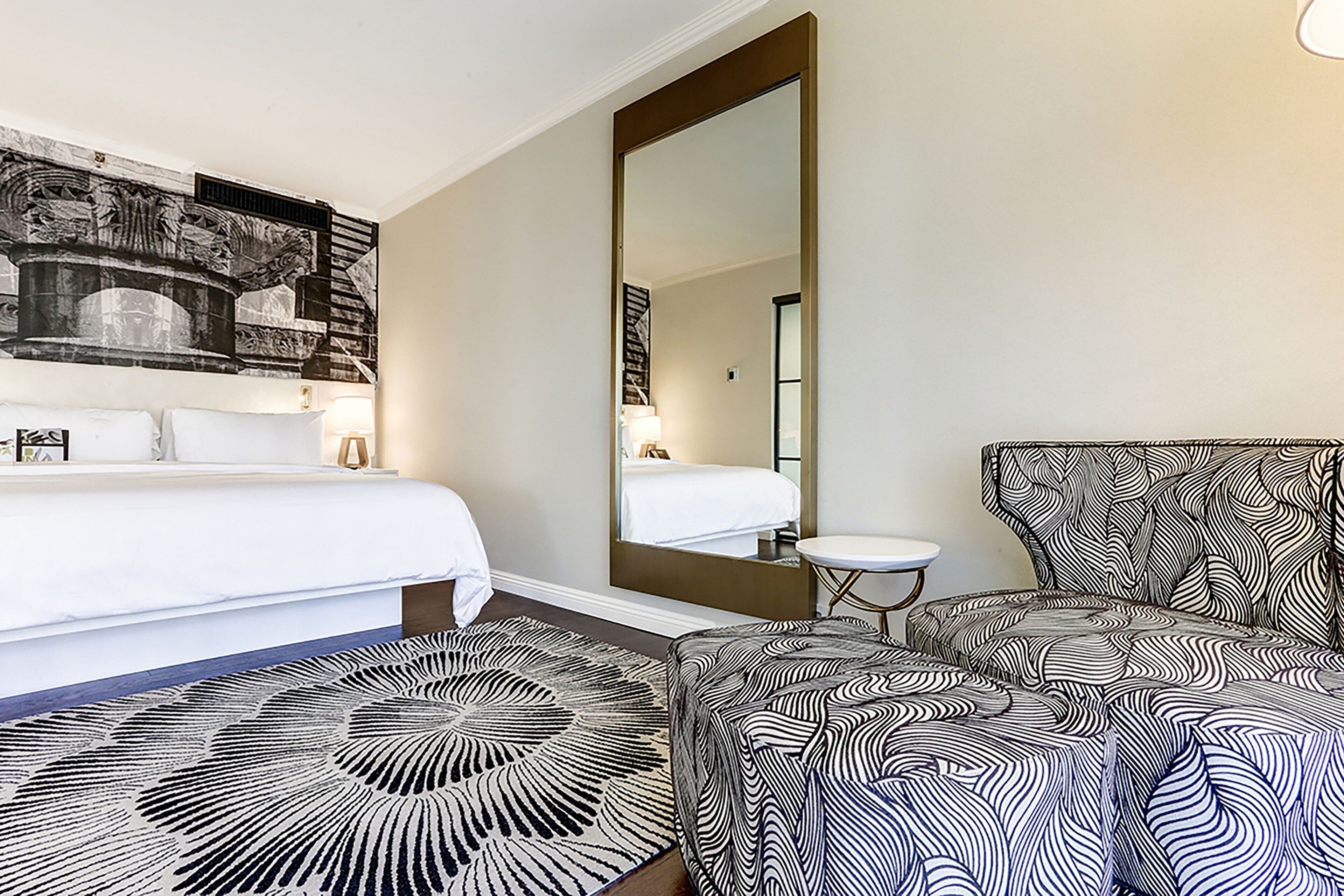 Hotellihuone, jossa on sänky, suuri peili, kuviollinen nojatuoli ja pieni pöytä.