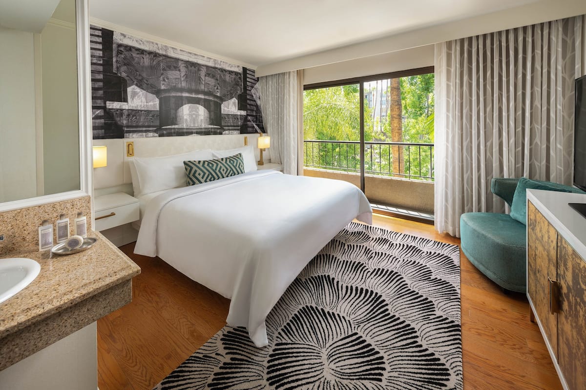 A hotel room with a large bed, a bathroom with a sink and mirror, a balcony with a view of greenery, and a TV on the wall.