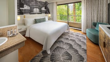 A hotel room with a large bed, a bathroom with a sink and mirror, a balcony with a view of greenery, and a TV on the wall.