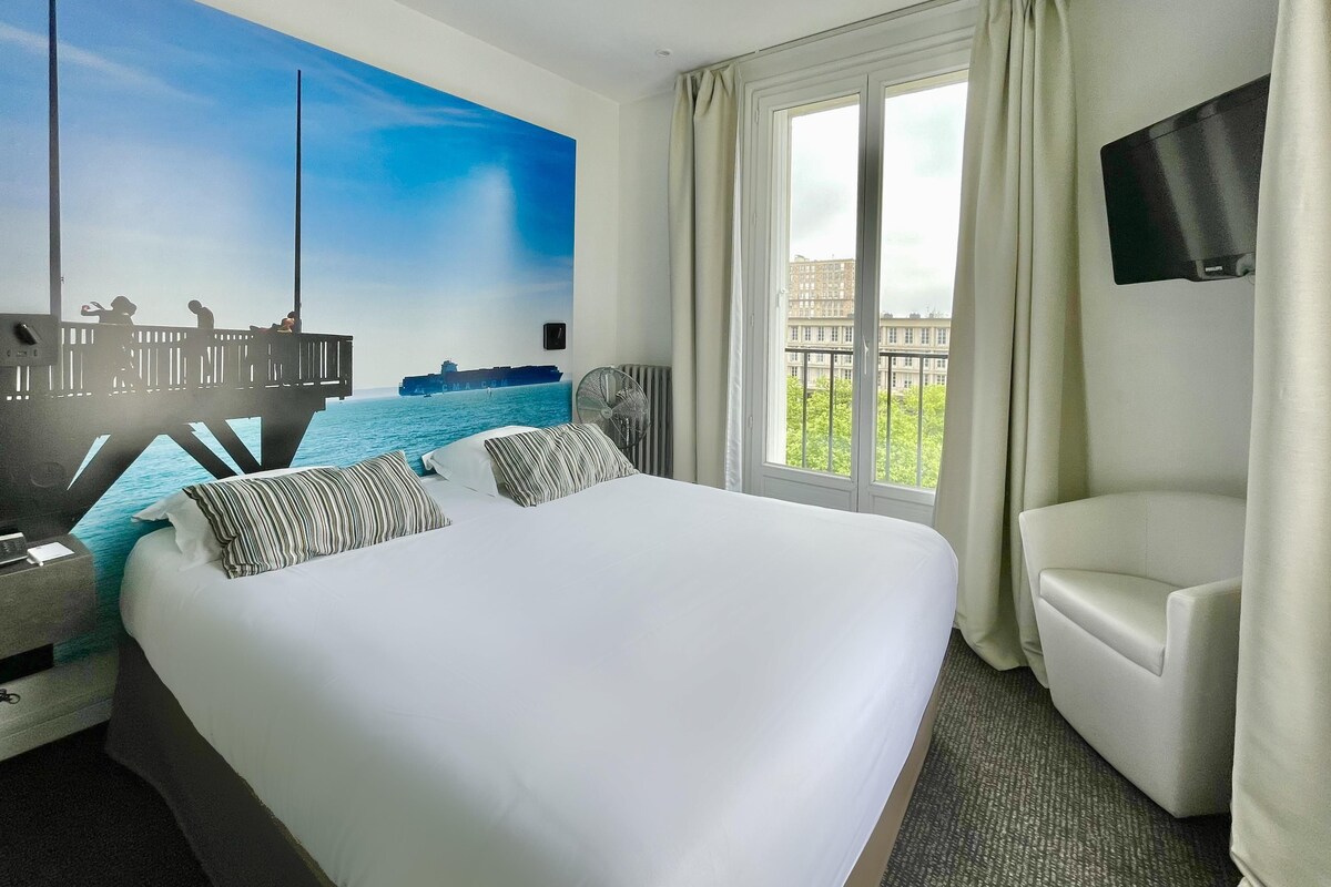 A hotel room with a large bed, a desk with a chair, a flat-screen TV, and a large window with a view of buildings.