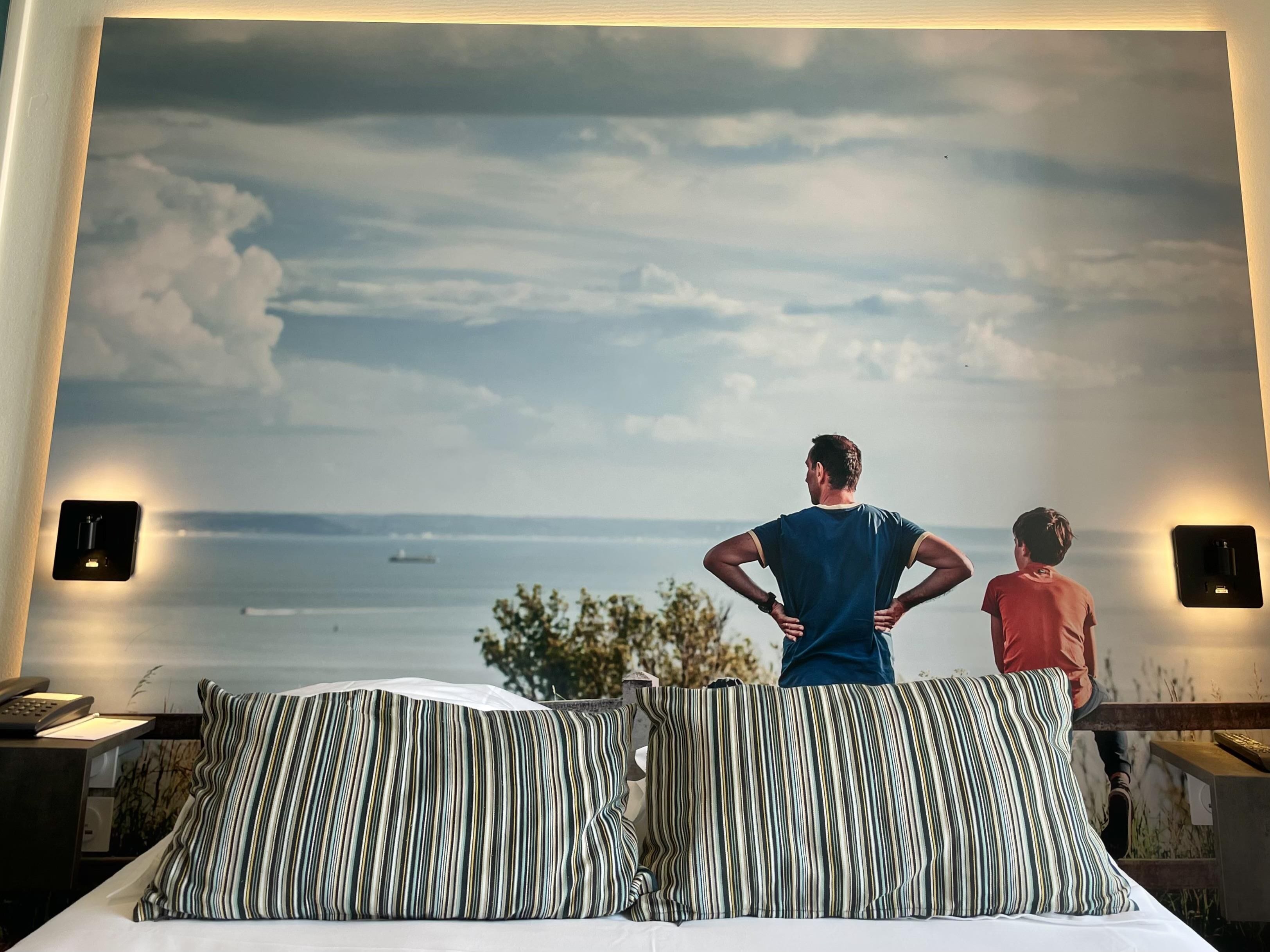 Une chambre avec une grande fresque murale représentant un paysage marin, un lit avec des oreillers à rayures et une table de chevet sur laquelle se trouvent un téléphone et une lampe.