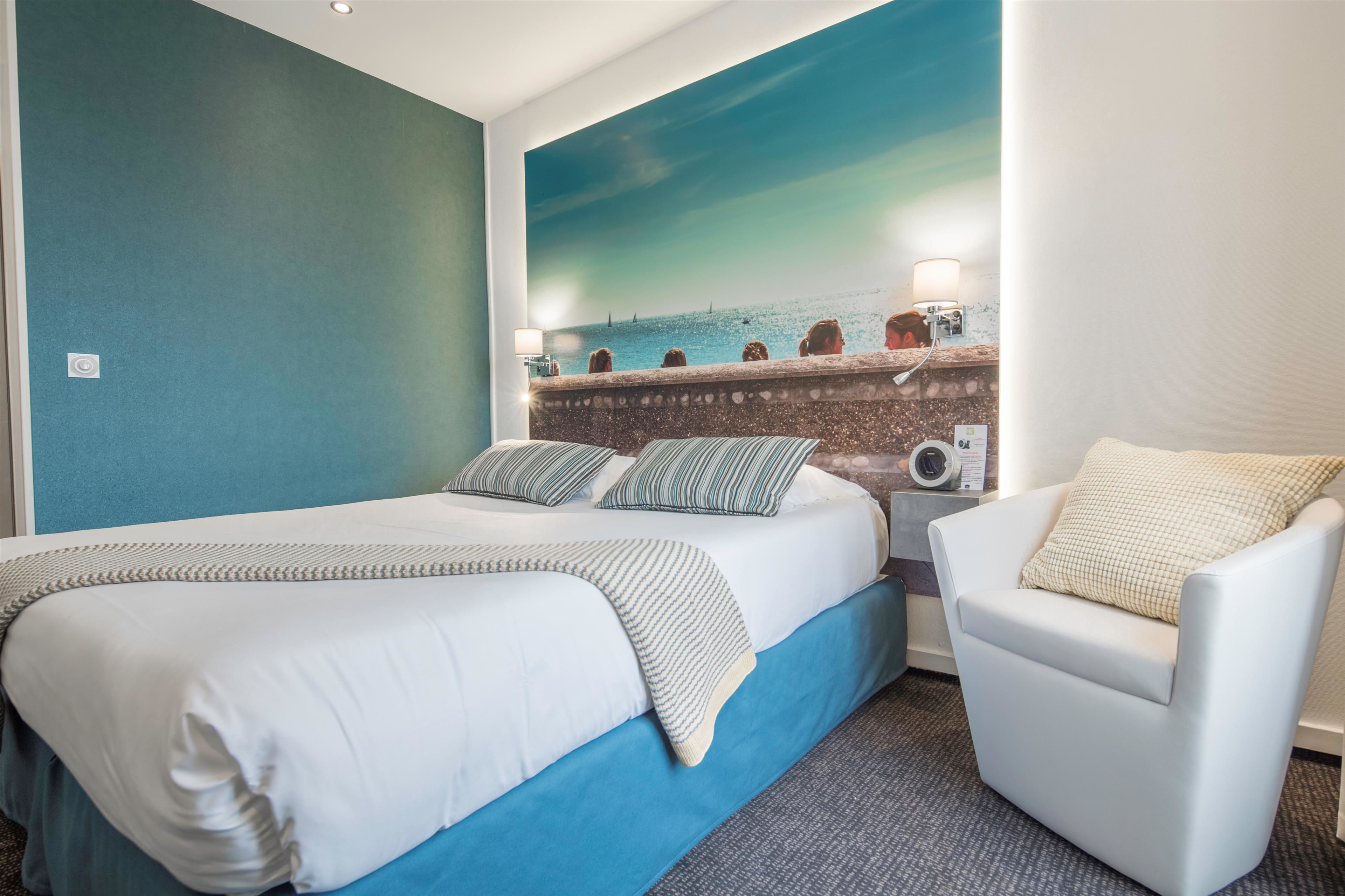 Une chambre d’hôtel avec un grand lit, une chaise, une fresque murale représentant un paysage marin et une table de chevet avec une lampe.