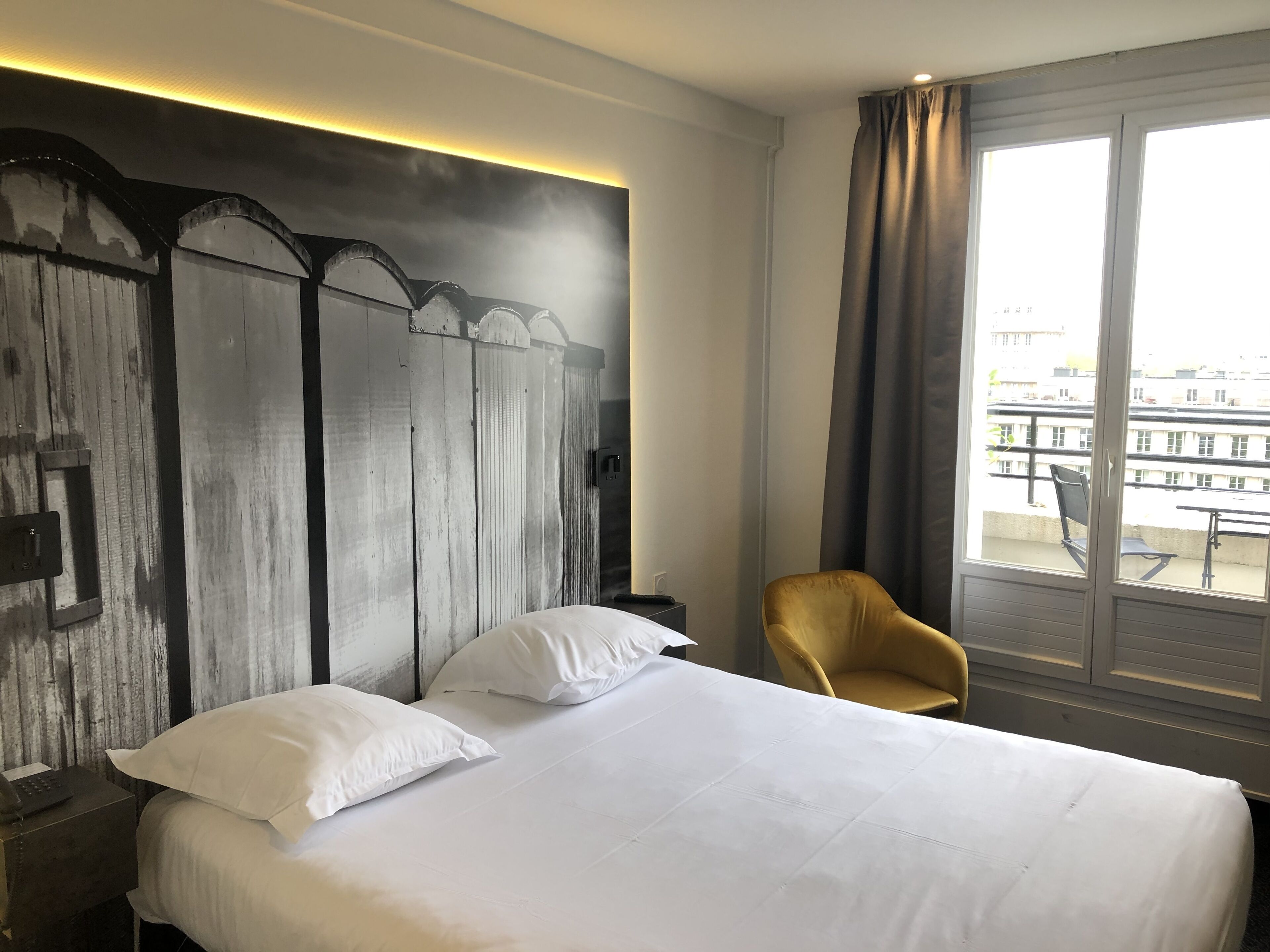 Una habitación de hotel con una cama grande, un cabecero de madera, una silla amarilla individual y vistas de la ciudad a través de un ventanal amplio.