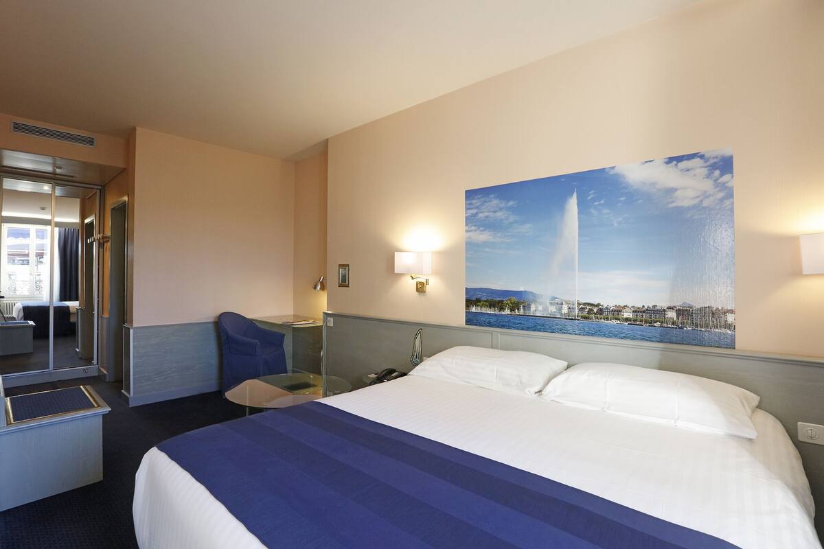 Quarto de hotel com cama, escrivaninha, cadeira, espelho e uma grande janela com vista para a cidade.