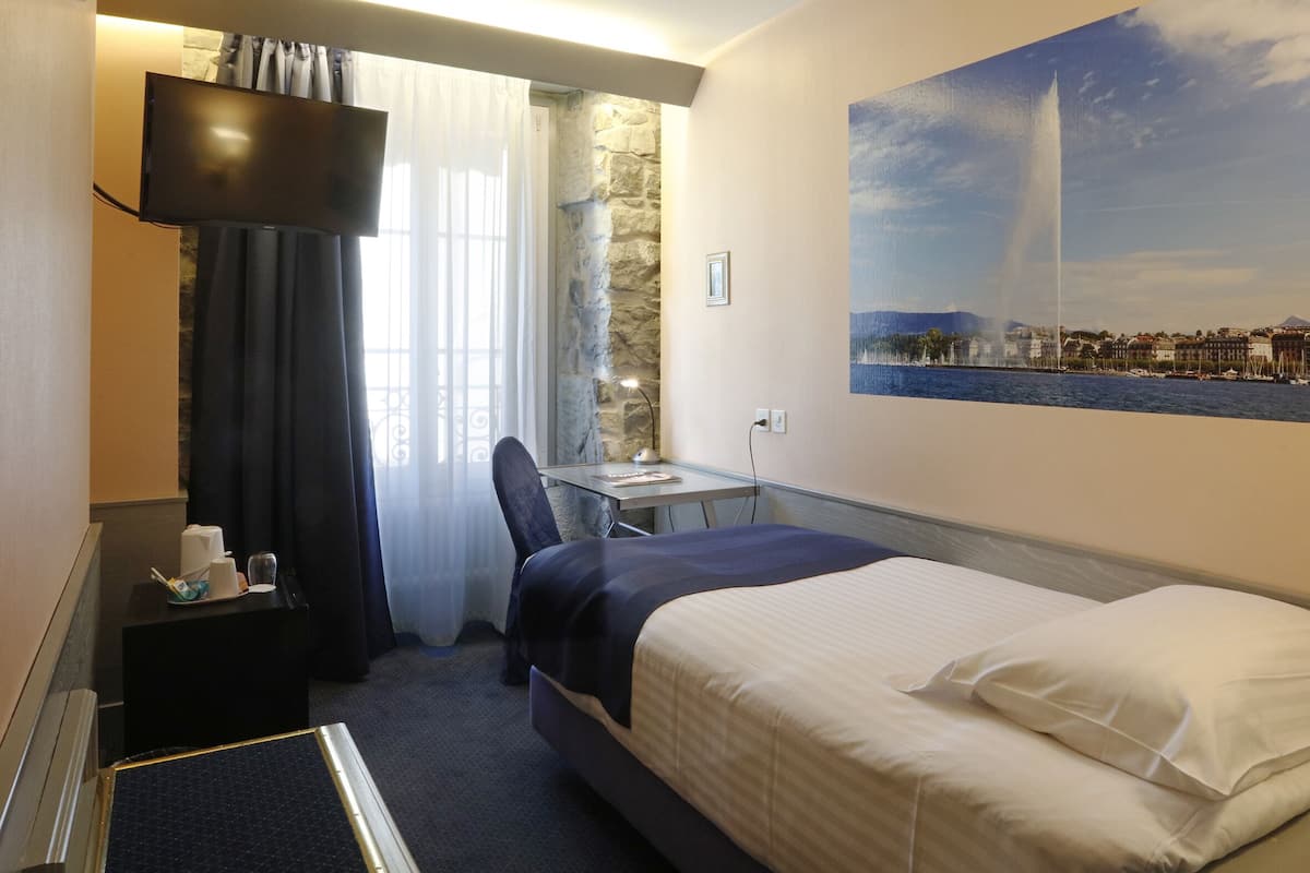 Quarto de hotel com cama, escrivaninha, cadeira, televisão, quadro de uma fonte e parede de pedra.