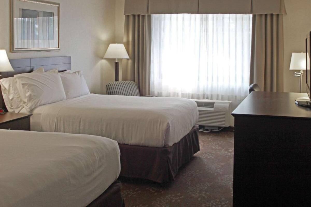 Une chambre d’hôtel avec deux lits, un bureau, une chaise, une lampe et une grande fenêtre avec des rideaux.