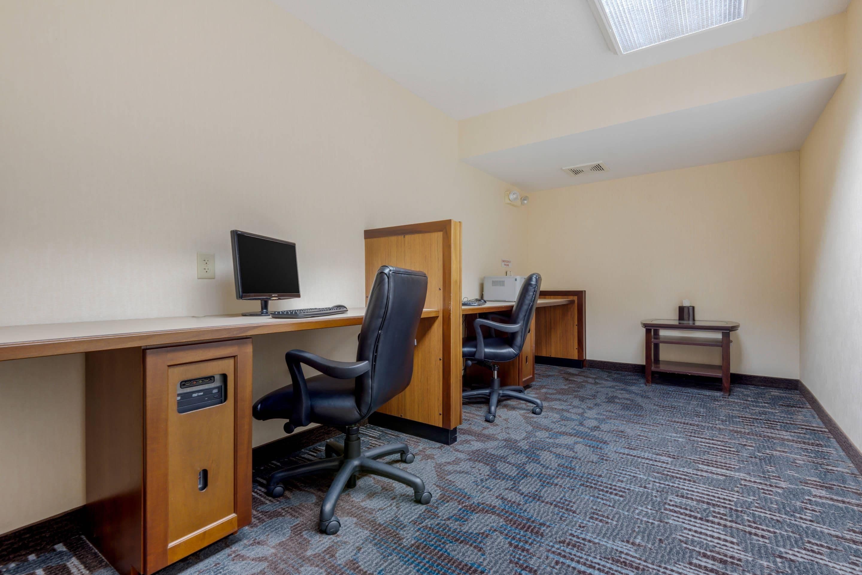 A hotel room with a desk, two chairs, a computer, a small table, and a carpeted floor.