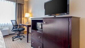A hotel room with a flat-screen TV mounted on a cabinet, a desk with a lamp, a chair, and a bed.