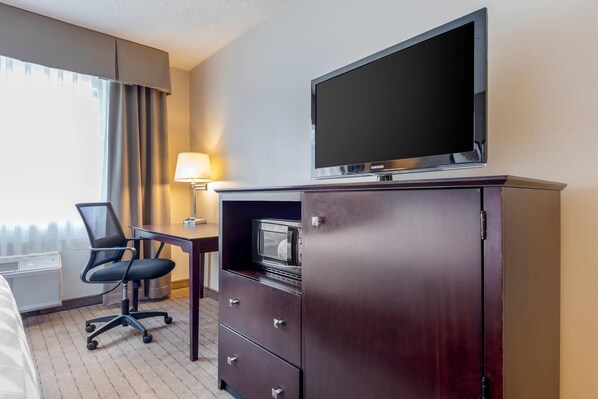 Ein Hotelzimmer mit Flachbildfernseher an einer Kommode, einem Schreibtisch mit Lampe, einem Sessel und einem Bett.