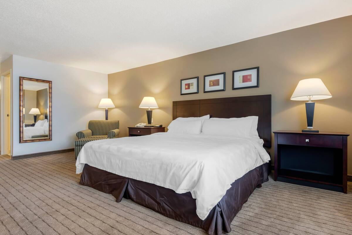 A hotel room with a large bed, two bedside tables with lamps, a chair, a mirror, and framed pictures on the wall.