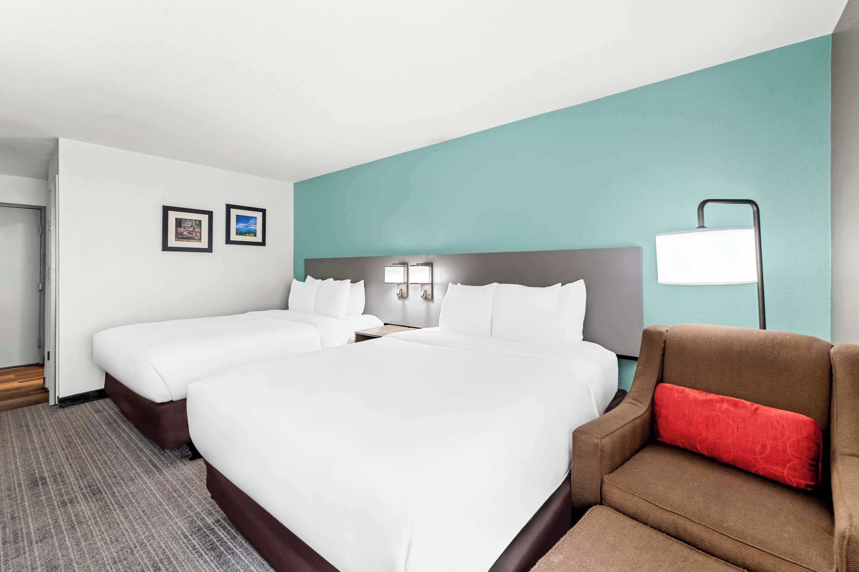 A hotel room with two beds, a brown armchair, a red pillow, a white lamp, and framed pictures on the wall.