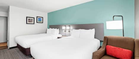 A hotel room with two beds, a brown armchair, a red pillow, a white lamp, and framed pictures on the wall.