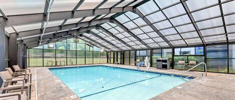 Une piscine intérieure dotée d’un toit en verre, de chaises longues et d’un espace aménagé au bord de la piscine avec une table et des chaises.